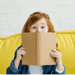 Red haired preschool girl holding book on a yellow couch
